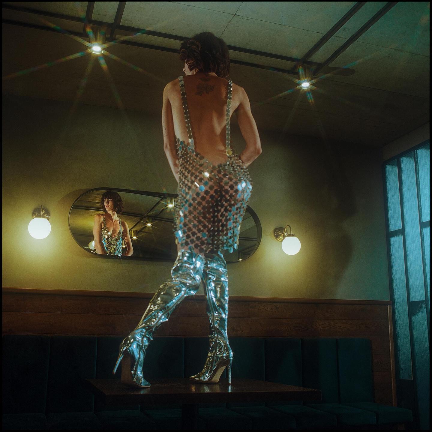 A person in a sparkly dress and boots stands on a table, with their reflection visible in a mirror.