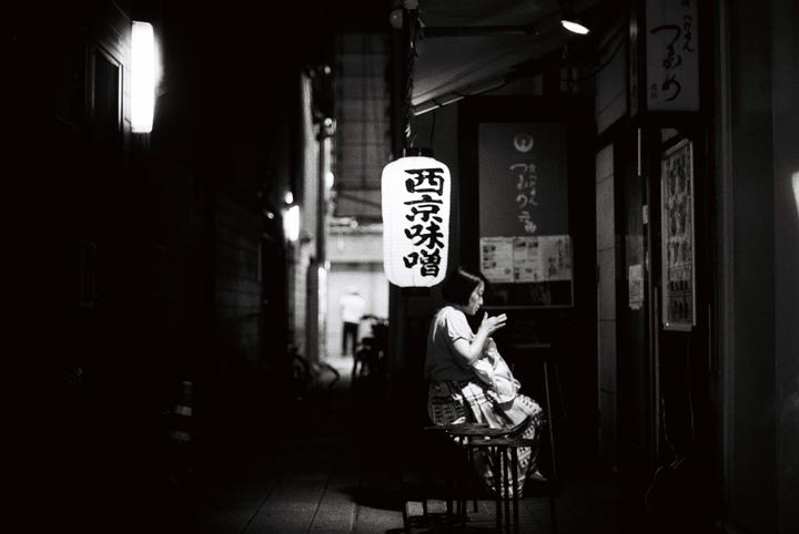 A woman sits on a stool under a hanging lantern on a dark street at night.