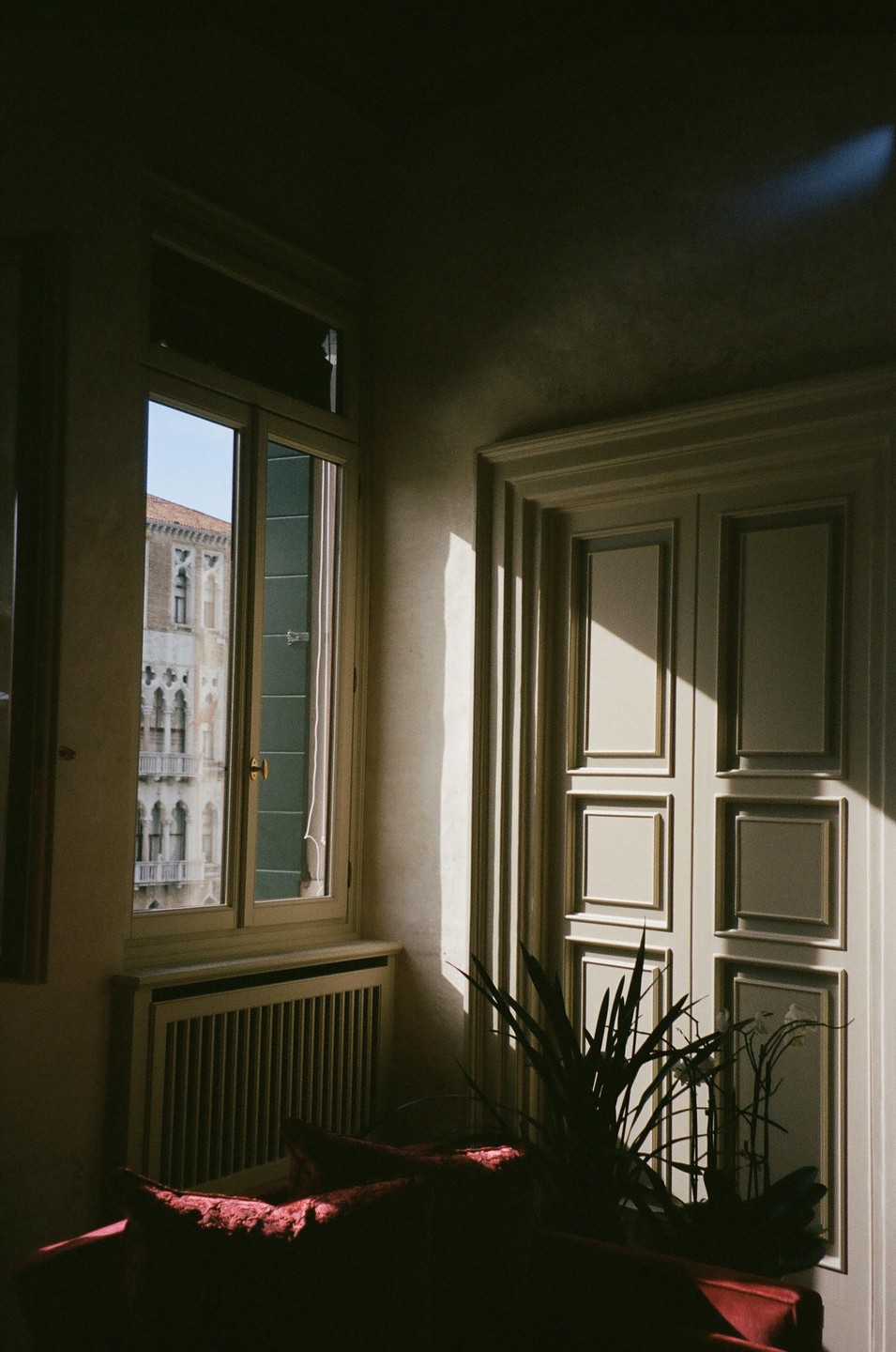 Sunlight streams through a window, illuminating part of a room with a plant and a red couch, while an old building is visible outside.