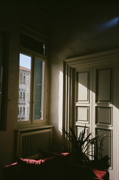 Sunlight streams through a window, illuminating part of a room with a plant and a red couch, while an old building is visible outside.