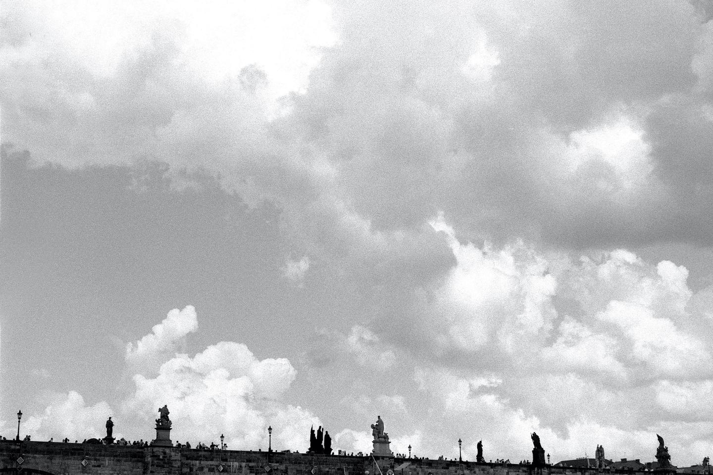 A low-angle view shows a bridge with statues and people against a cloudy sky.