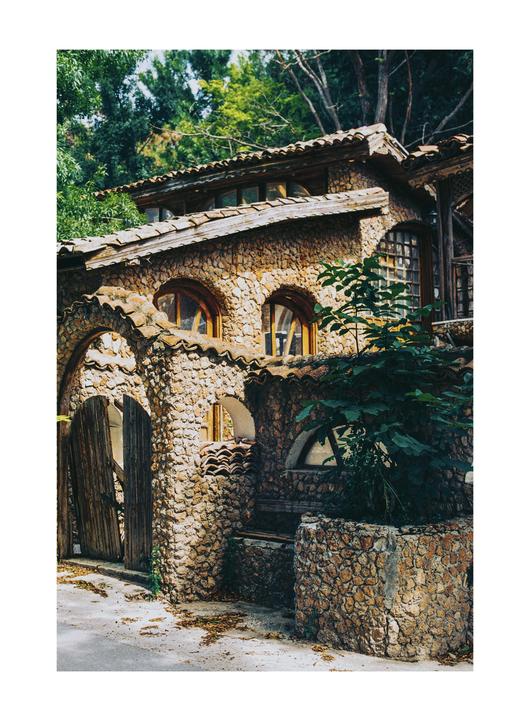 A stone house with a tiled roof and arched windows is surrounded by green foliage.
