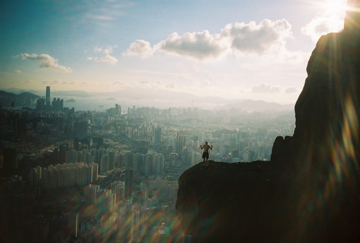 A man stands on a rocky outcrop overlooking a sprawling city with a hazy sky and lens flare.