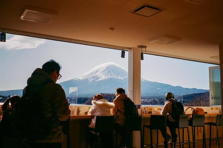 People sit at a counter looking out a window at a snow-capped mountain.