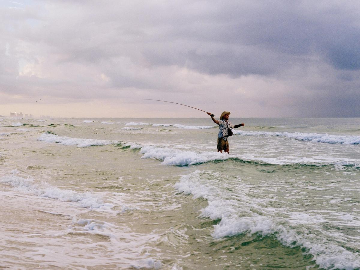 A man wearing a cowboy hat stands in the ocean holding a fishing rod as waves crash around him under a cloudy sky.