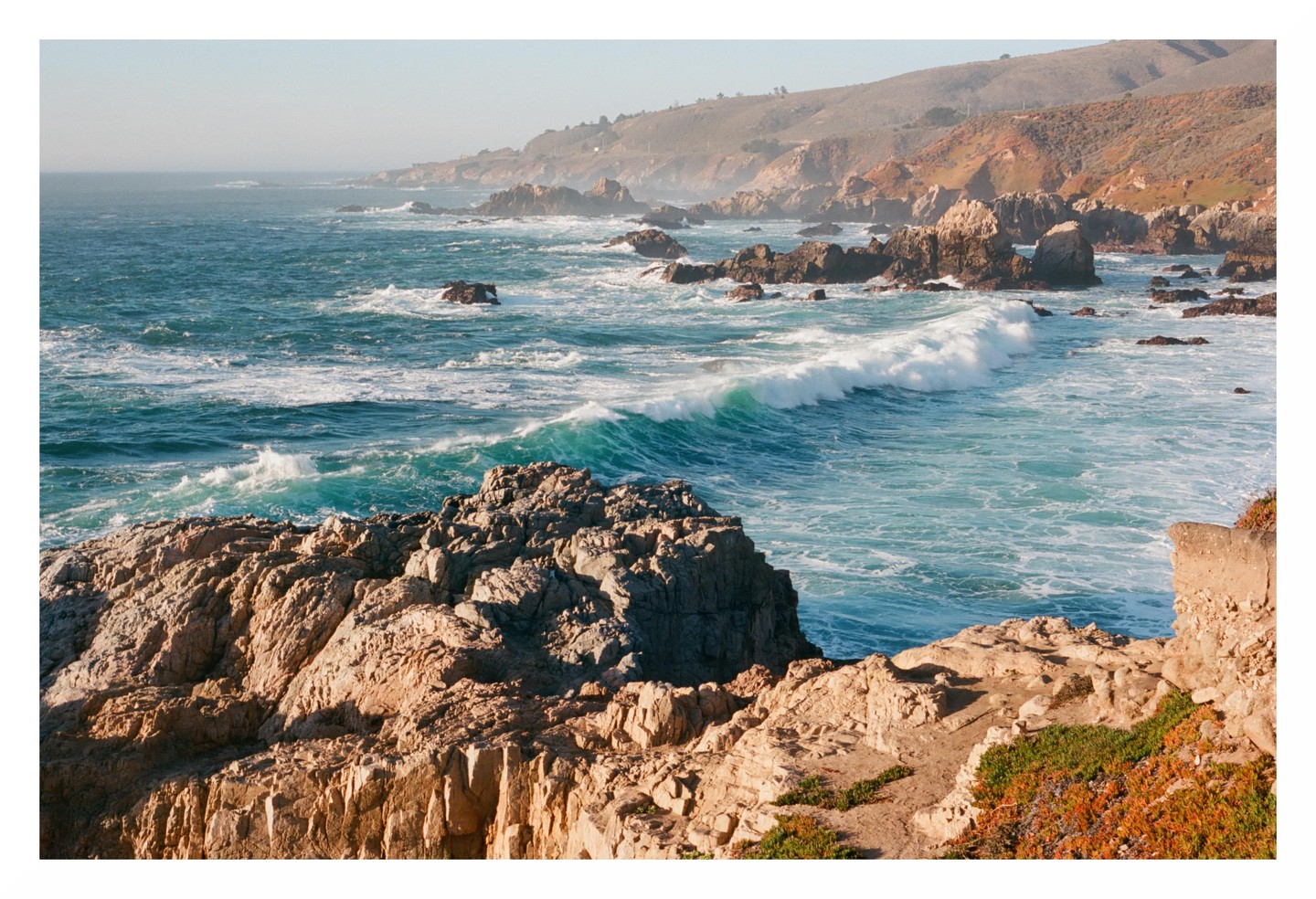 Waves crash against a rocky coastline with a hazy sky and distant hills.