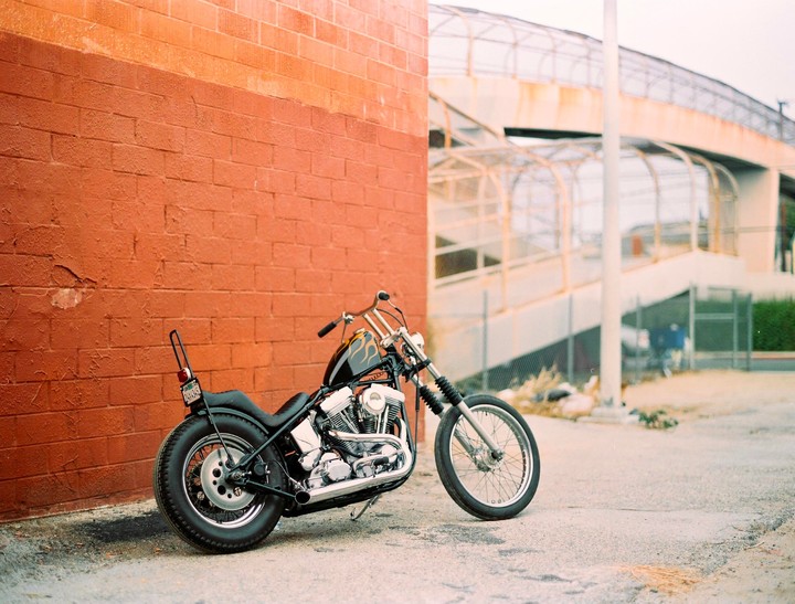A black motorcycle with chrome details is parked next to an orange brick wall, with an elevated walkway in the background.