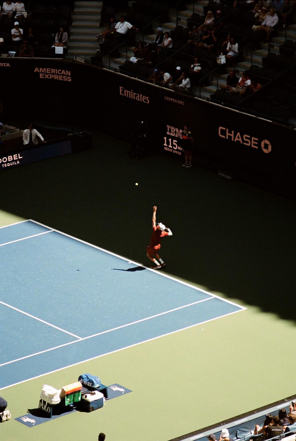 A tennis player in a red shirt serves a tennis ball on a court with spectators in the stands.