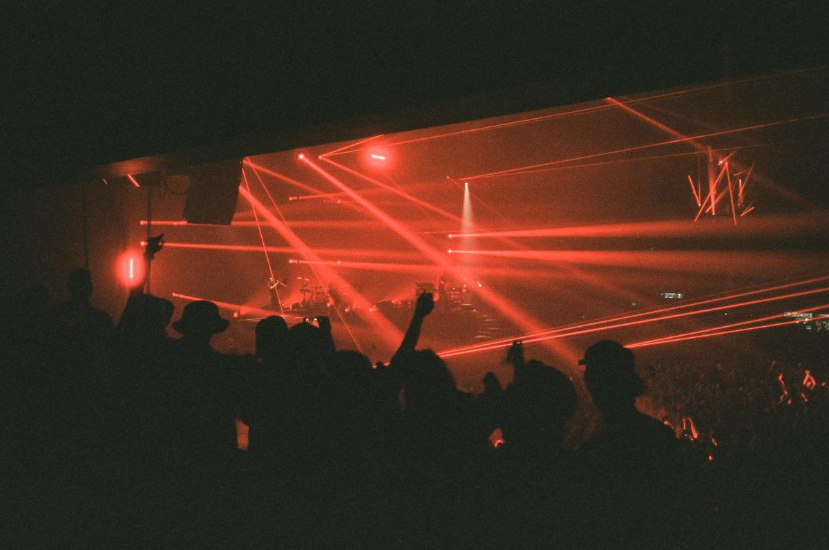 A crowd of people are silhouetted against a stage illuminated by red laser lights at a concert.
