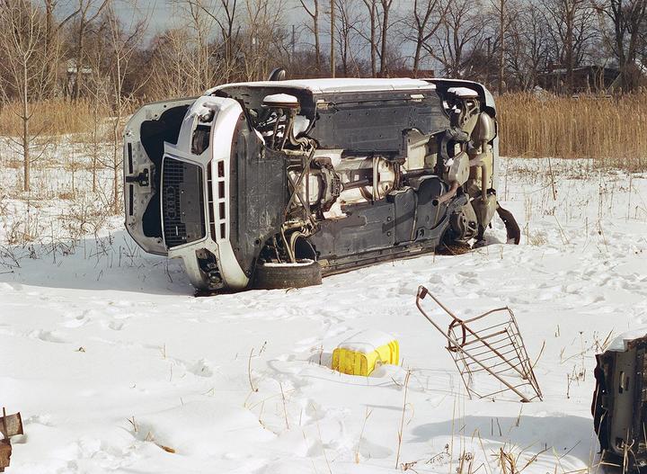 A white car lies overturned in a snowy field next to a shopping cart and a yellow container.