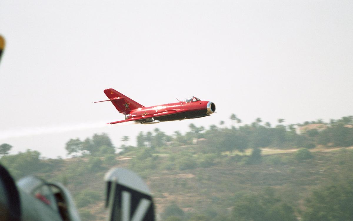 A red jet flies through the air with a trail of smoke behind it, above a blurred landscape of green trees.