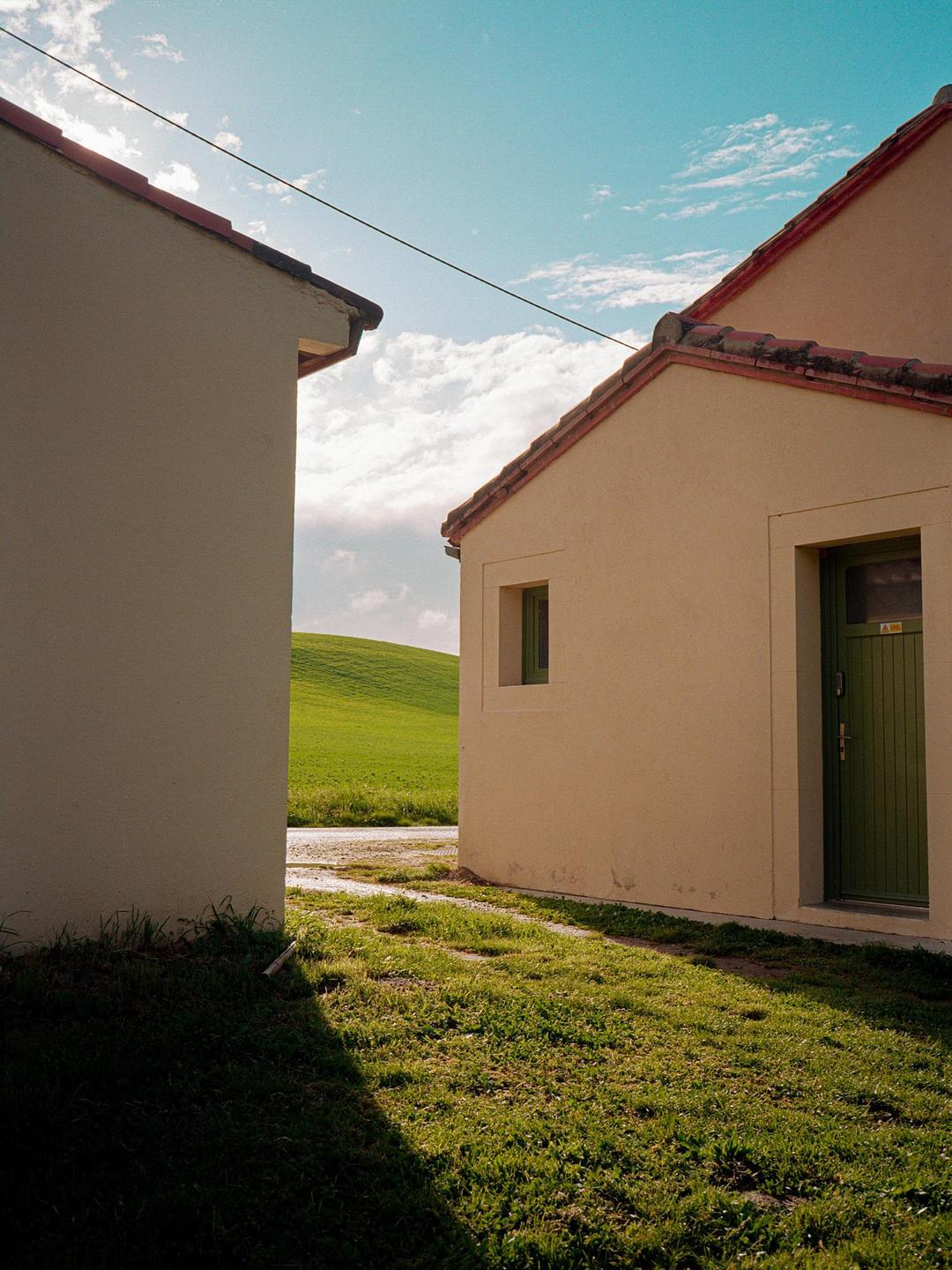 Two buildings with tiled roofs flank a grassy path leading to a bright green hill under a blue sky with clouds.