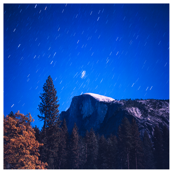 A long exposure photograph captures star trails above Half Dome and a forest at night.
