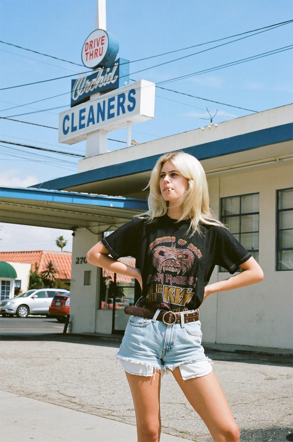 A woman stands in front of a drive-thru cleaners sign under a clear sky.