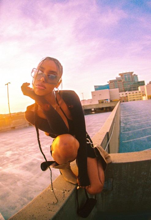A woman in sunglasses crouches on a concrete ledge with a city skyline in the background at sunset.