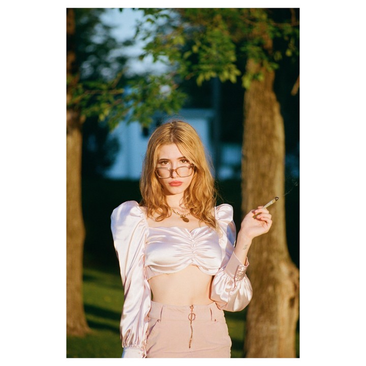 A woman with glasses and a shiny pink top holds a lit cigarette in a park.