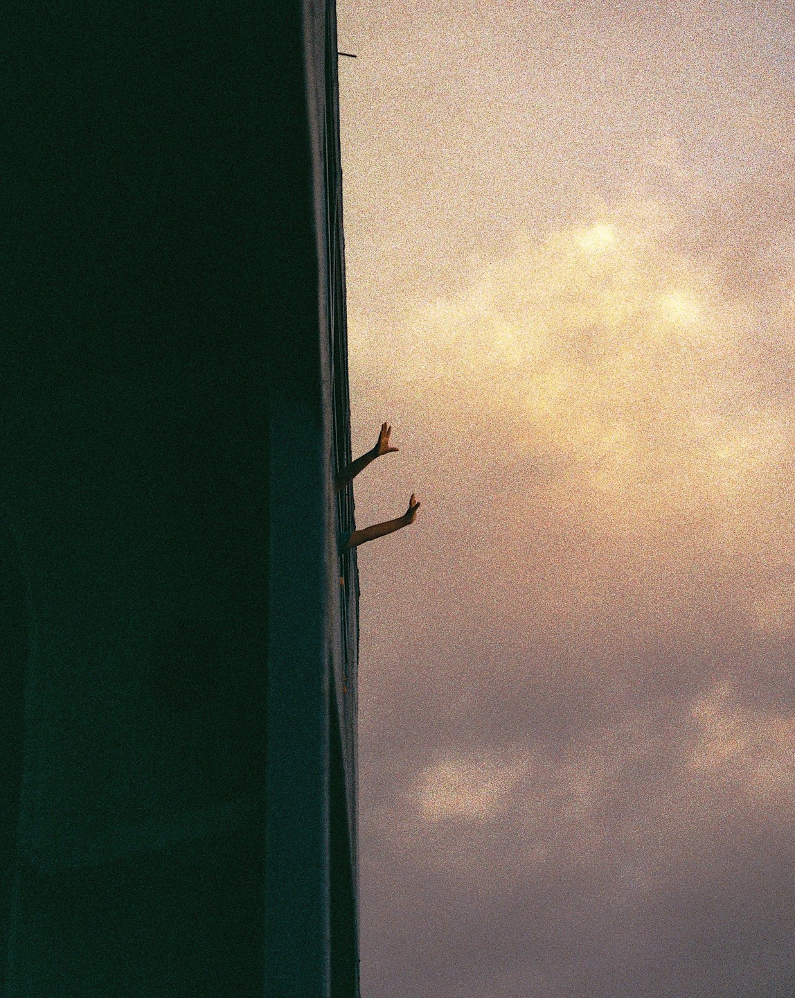 Two hands reach out from a narrow opening in a building against a cloudy sky.