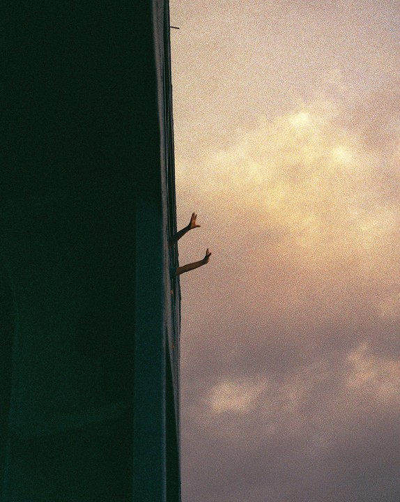 Two hands reach out from a narrow opening in a building against a cloudy sky.