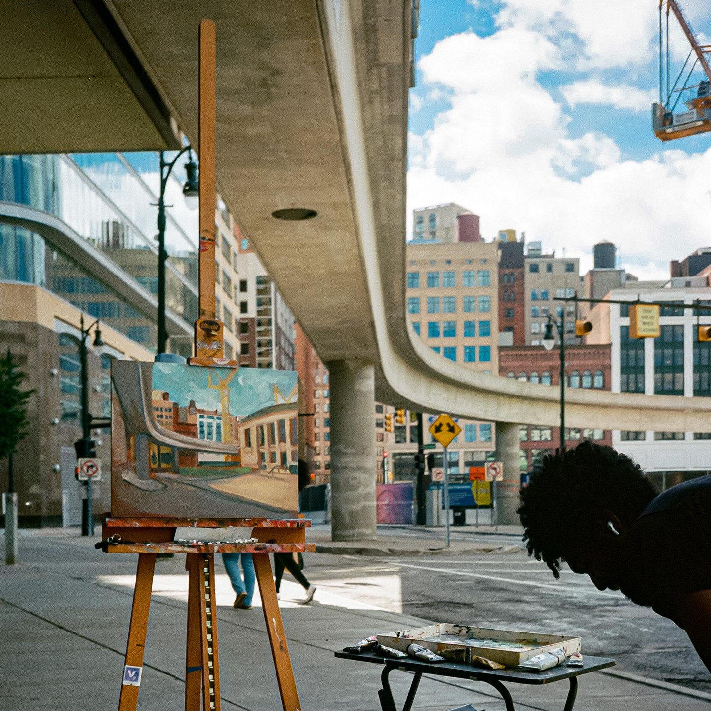A man paints a city street scene on a canvas set up on a sidewalk next to an elevated highway.