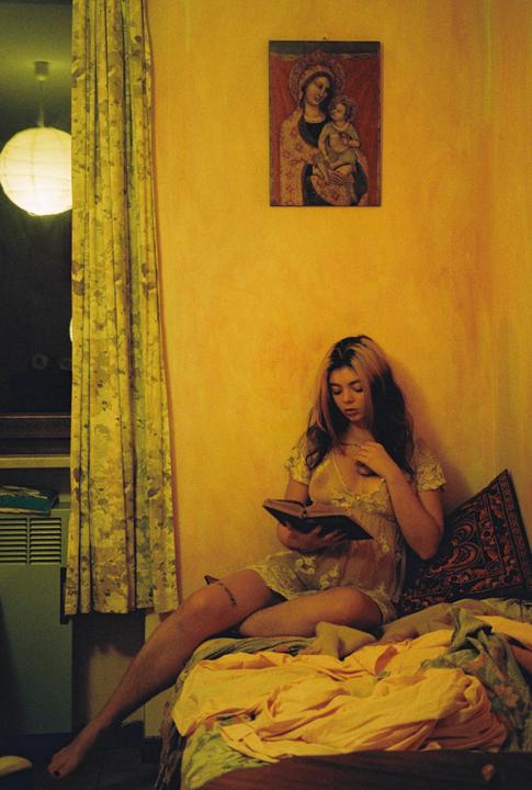 A woman sits on a bed reading a book next to a window with a paper lantern and a religious painting on the wall.