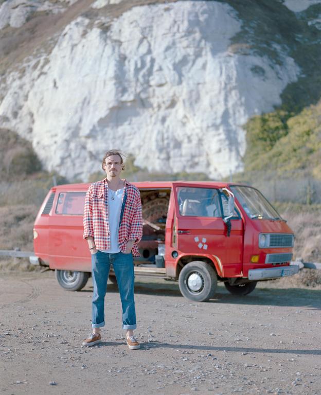 A man stands in front of a red van with a white cliff in the background.