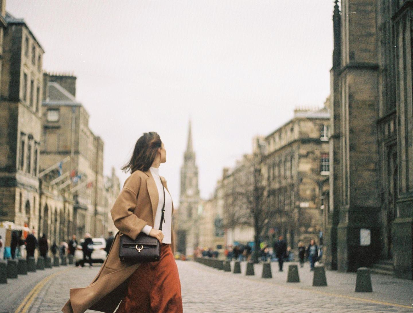 A woman in a long coat and skirt walks down a cobblestone street lined with historic buildings, with a prominent spire in the background.