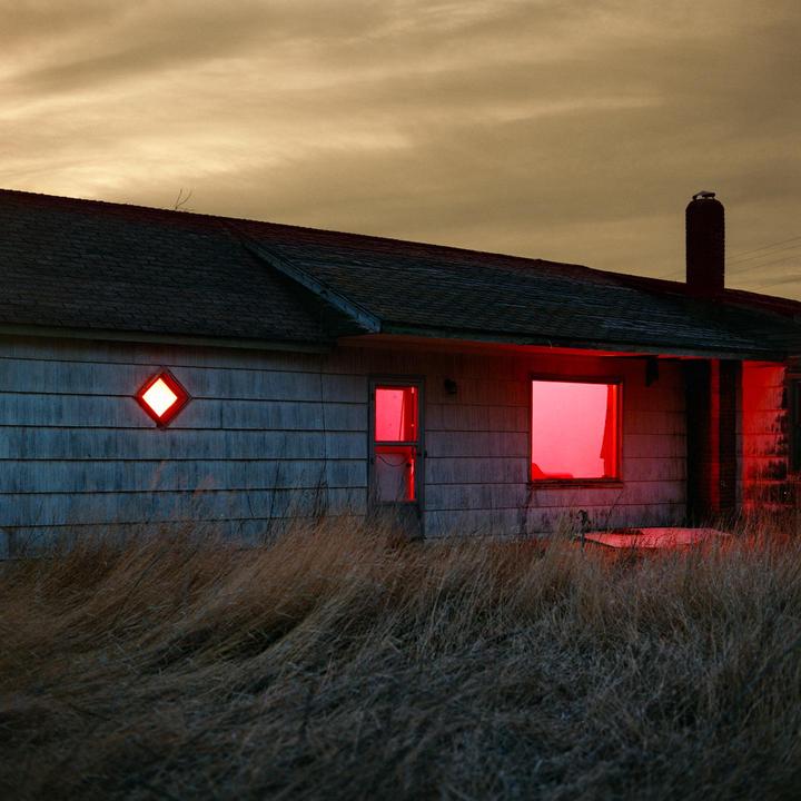 An abandoned house with glowing red windows stands in a field of tall, dry grass under a cloudy sky.