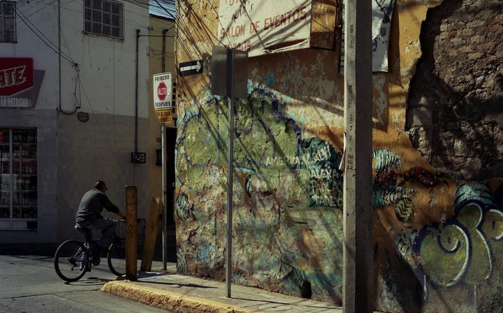 A man rides a bicycle past a building with peeling paint and graffiti on a sunny street.