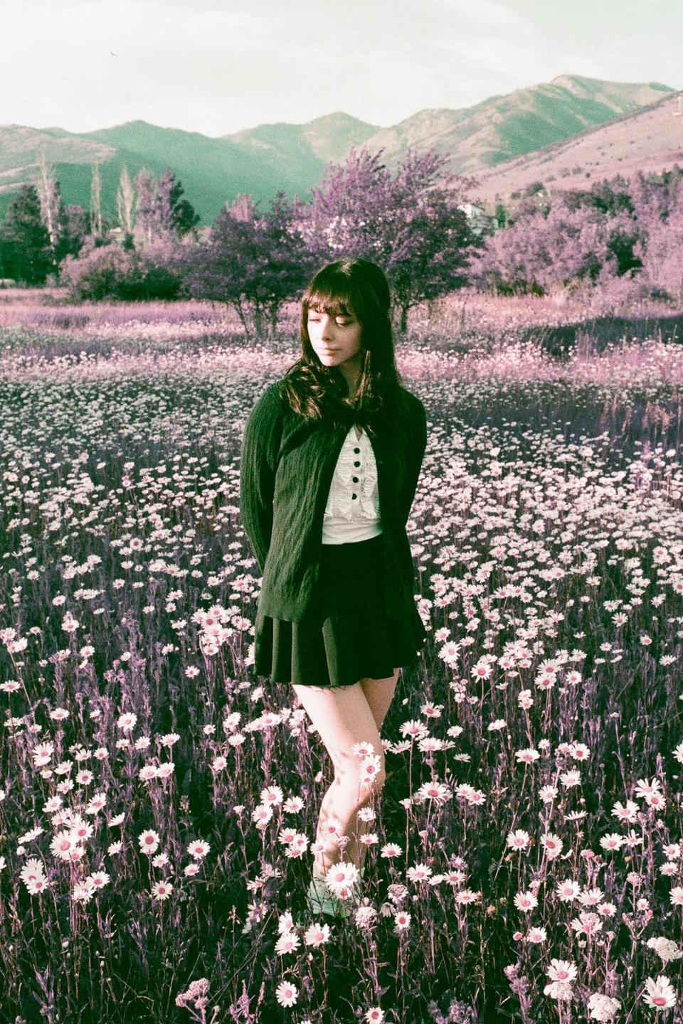 A woman stands in a field of white flowers with purple foliage and mountains in the background.