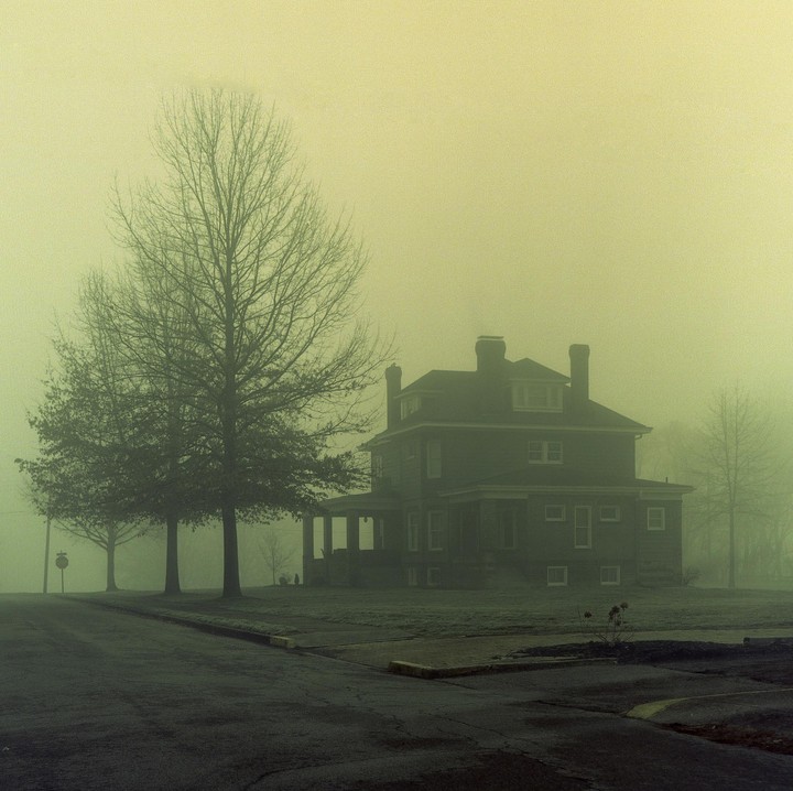 A large house with a porch stands next to a street and trees in a thick fog.
