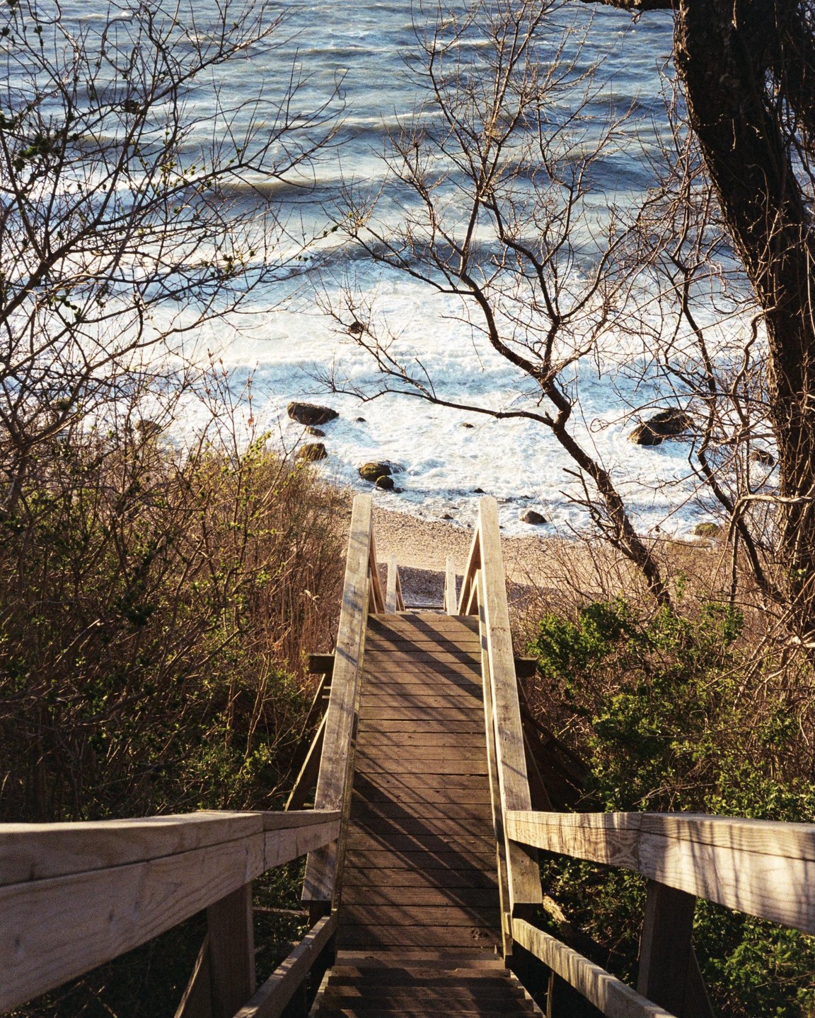 A wooden staircase leads down to a rocky beach with waves crashing on the shore.