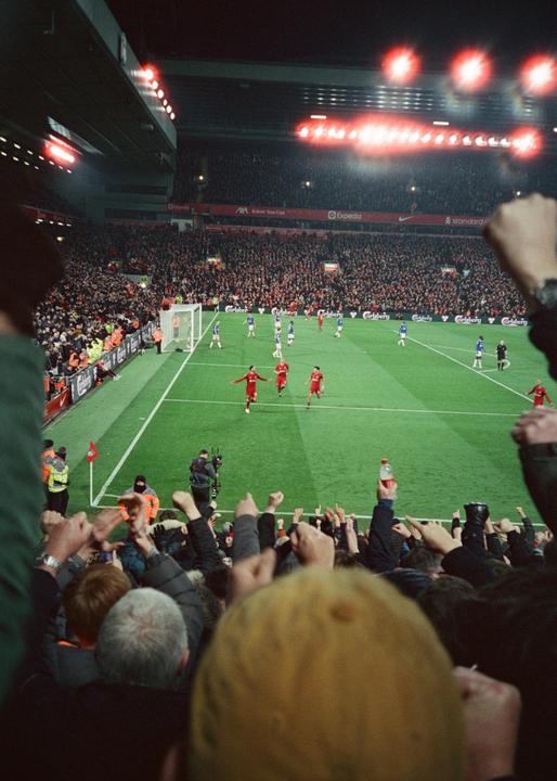 A crowd of people with their arms raised watches a soccer game from the stands at night.
