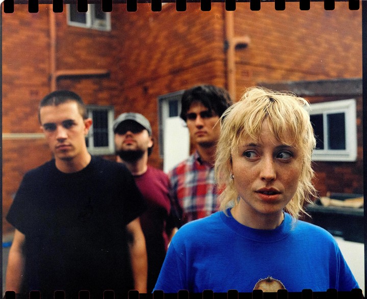A woman with blonde hair looks to the right while three men stand behind her.