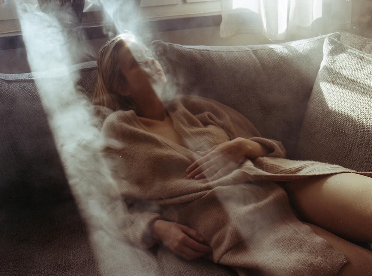 A woman reclines on a sofa, enveloped in a hazy, smoky atmosphere with light streaming in from a window.