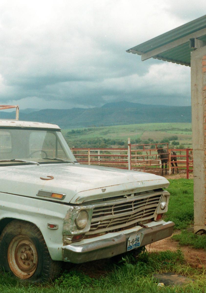 A light blue pickup truck is parked in a grassy area next to a fence with a horse behind it, with mountains in the background under a cloudy sky.