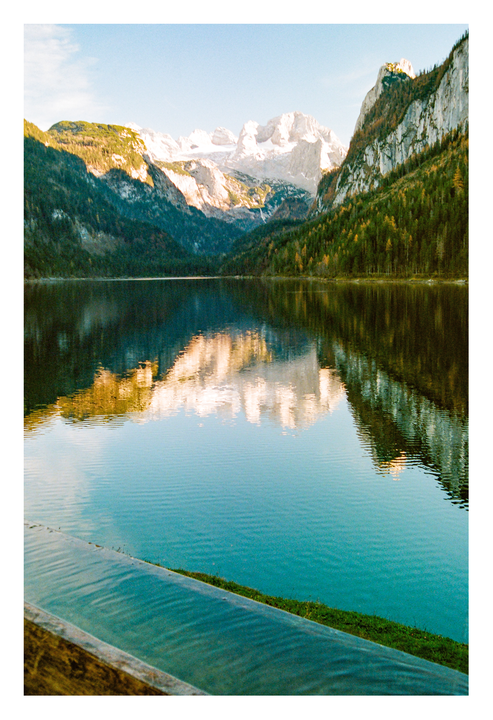 A lake reflects snow-capped mountains and a forest under a clear sky.