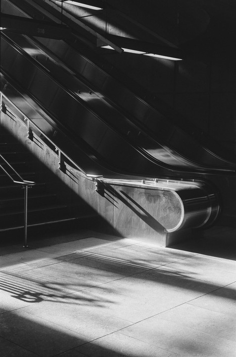 Sunlight casts strong shadows across an escalator and a tiled floor.