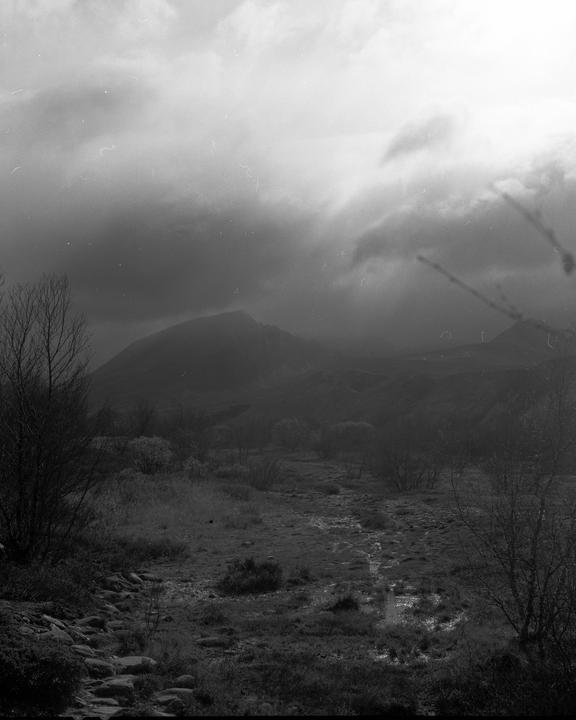 A dark mountain rises under a cloudy sky, with a stream and bare trees in the foreground.