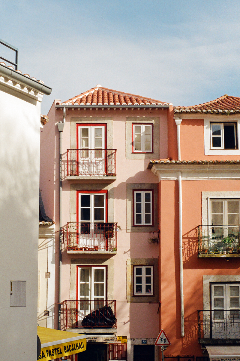 A light pink building with red-framed windows and balconies stands next to an orange building under a clear sky.