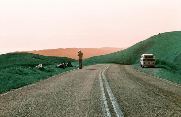 A person stands on a road next to a parked van, with green hills and a pink sky in the background.