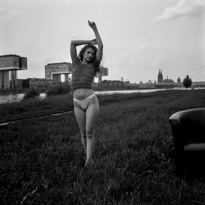 A woman stands in a grassy field with her arms raised, with a city skyline and river in the background.