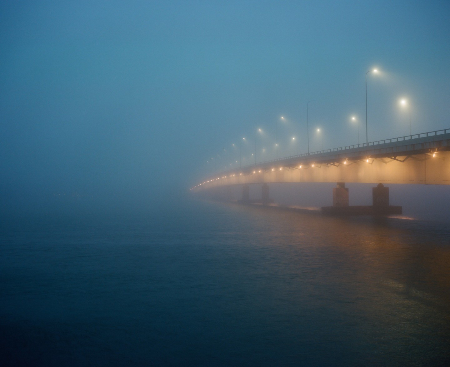 A long bridge with many streetlights extends into the distance over water, shrouded in fog.
