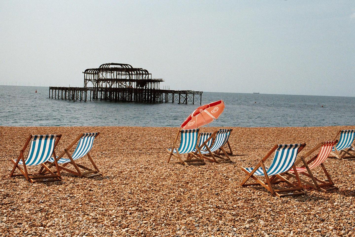 Several striped deck chairs and a red umbrella sit on a pebble beach with a ruined pier in the background.