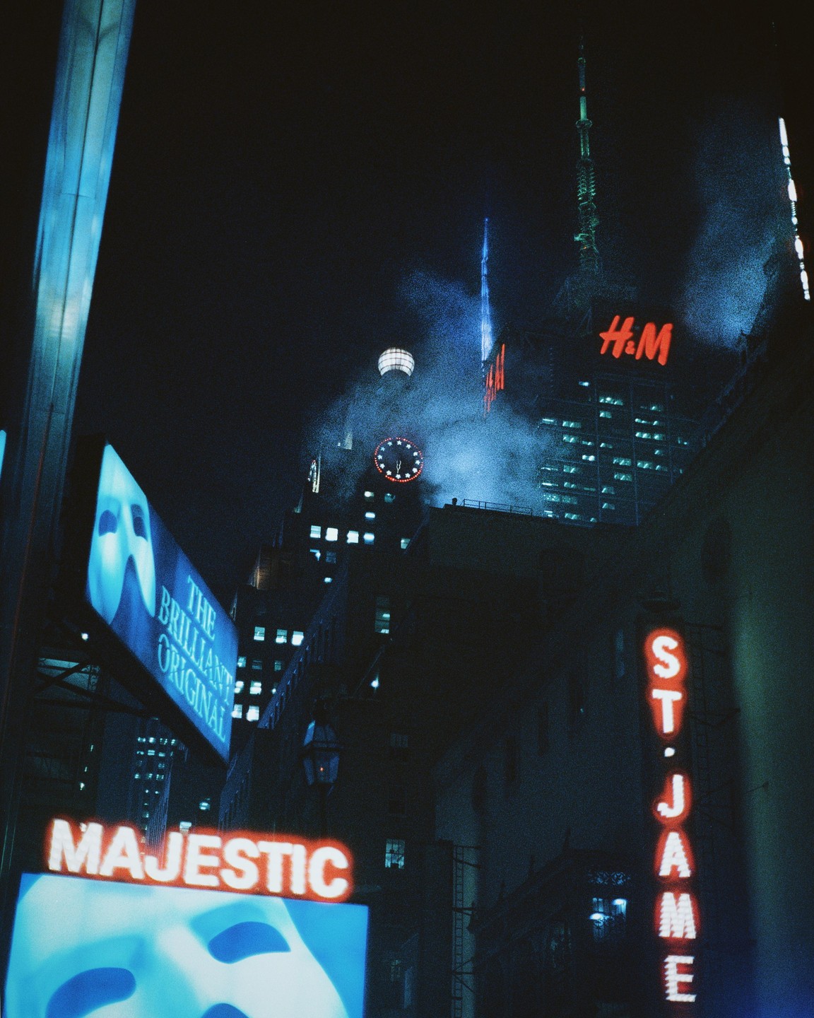 Neon signs illuminate a city street at night, with a tall building and a clock visible in the background.