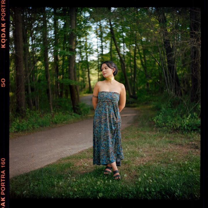 A woman in a strapless dress stands on a grassy path in a forest.