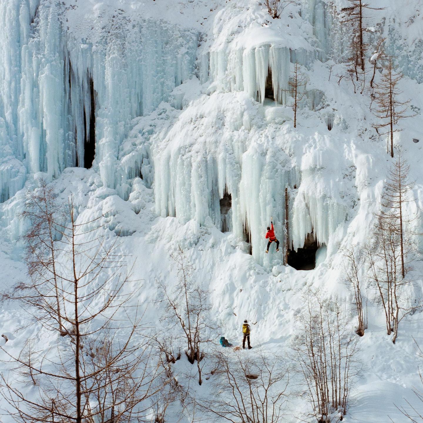 A person in red is ice climbing on a large frozen waterfall while another person stands below in a snowy landscape with bare trees.