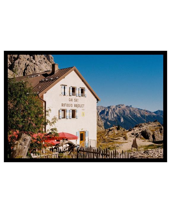 A white mountain hut with a red umbrella and a wooden fence stands against a backdrop of rocky mountains and a clear blue sky.