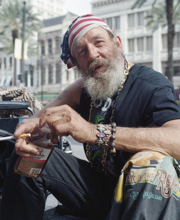 A man with a white beard and an American flag bandana smiles while holding a bottle of whiskey and a cigarette.