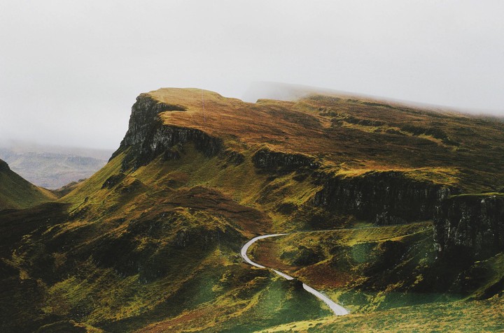 A winding road cuts through a vast, rugged landscape of green and brown hills under a hazy sky.