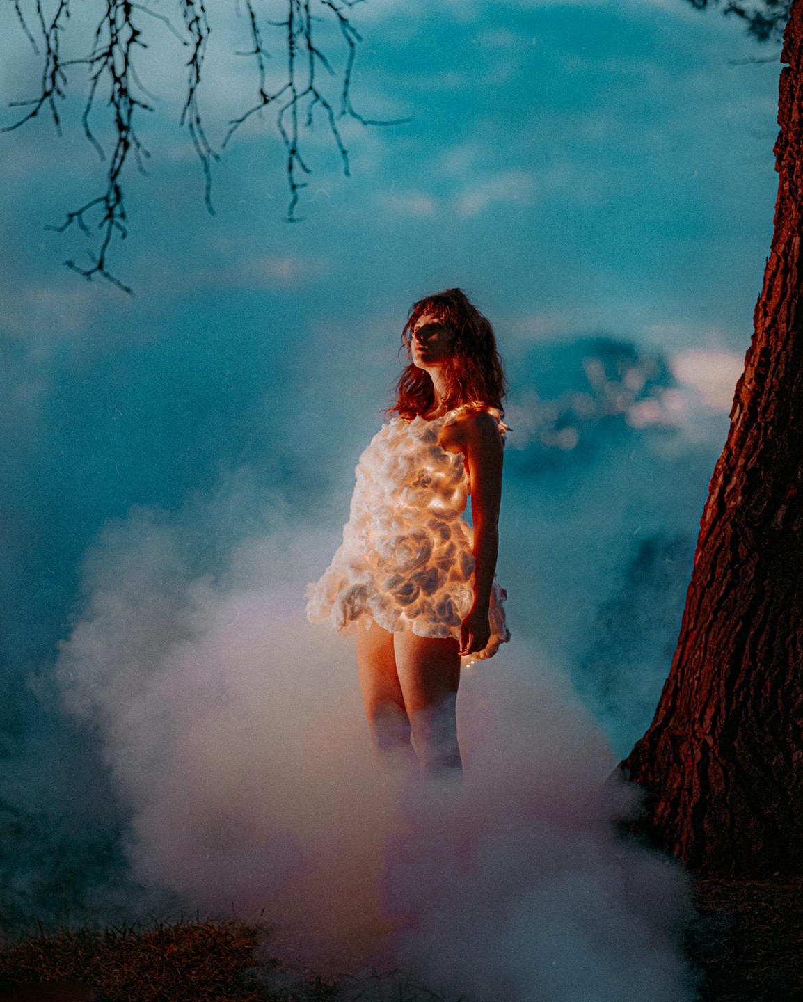 A woman in a glowing cloud-like dress stands in smoke next to a tree trunk under a blue sky.